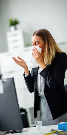 Young businesswoman with allergy symptoms blowing nose into paper tissue while working in a modern office, experiencing discomfort from cold or flu and needing to take a breakの素材