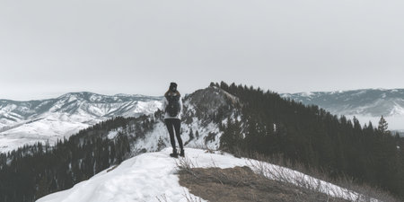 Woman standing on a snow-covered mountain ridge, looking out at vast snow-capped peaks and forests under an overcast sky, embodying solitude and a sense of adventure in natureの素材