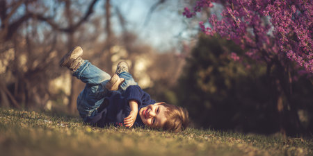 Young boy rolling and laughing on lush green grass in a park with pink blossoms, enjoying carefree spring playtime and bright sunny outdoors, pure childhood joy and motionの素材