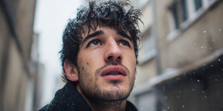 Young man standing outdoors during a winter day, having a pensive moment while looking into the falling snow, flakes settling on his hair and coat, reflecting urban coldnessの素材