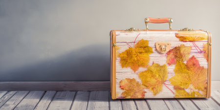 Vintage style suitcase standing on a light wooden floor with a gray wall background, its surface decorated with colorful autumn maple leaves, symbolizing seasonal travel and fall journeysの素材