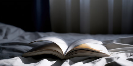Open book resting on a rumpled white duvet, illuminated by a beam of natural sunlight creating contrasting shadows and highlighting a quiet morning reading atmosphereの素材