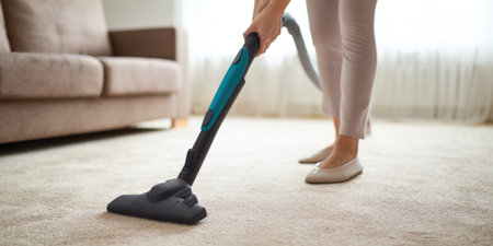 Woman vacuuming light-colored carpet in a modern living room, doing routine housework to maintain hygiene and tidiness; casual indoor cleaning activity and floor careの素材