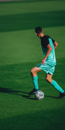 Male soccer player actively dribbling a football during a training session or match, wearing a light blue and black uniform on a vibrant green sports fieldの素材