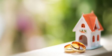 Golden wedding rings sitting next to a miniature house, symbolizing real estate, homeownership, commitment, family planning, love, and the future dreams of a coupleの素材