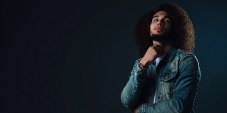 Young man with curly hair wearing a denim jacket standing against a dark background, looking up thoughtfully while contemplating future plans and opportunitiesの素材