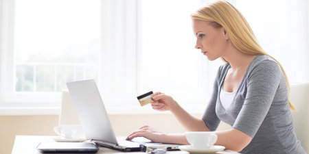 Blonde woman holding credit card and typing on laptop in bright living room, making secure online purchases from home, enjoying convenient e-commerce shopping and digital paymentsの素材
