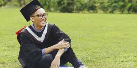 Graduating student celebrating academic achievement, sitting on green lawn outdoors, happily laughing and looking away, wearing a black cap and gown, symbolizing success and future opportunitiesの素材