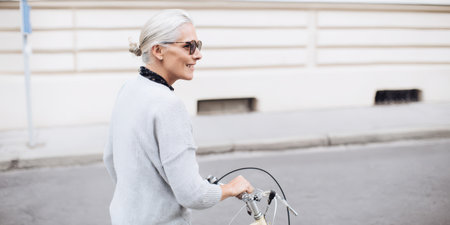 Senior woman with gray hair and sunglasses smiling as she walks her bicycle along a city street, enjoying an active, healthy urban retirement and carefree outdoor lifestyleの素材