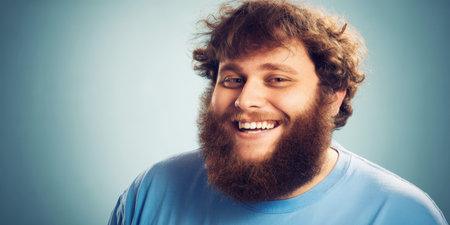 Happy young man with messy curly hair and a long beard smiling broadly, radiating joy, confidence and authenticity against a clean blue studio background with copy spaceの素材