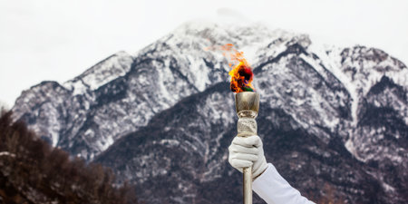 Hand in white glove holding olympic torch with bright flame against snow-capped mountain peaks and cold winter sky, symbolizing hope, achievement and the spirit of the gamesの素材
