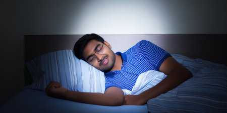 Young indian man sleeping soundly on his side in bed, resting comfortably under a duvet in a dark bedroom, enjoying peaceful slumber and good quality sleepの素材
