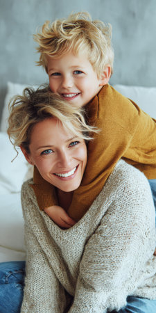 Happy blonde mother smiling while embracing her young son from behind, boy smiling and resting head on mother's shoulders, sharing a moment of family love and affectionの素材