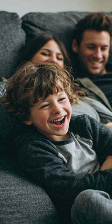 Young boy laughing on a gray sofa, relaxing with parents blurred behind him, sharing a cozy, joyful family moment of playful bonding and warmth at homeの素材