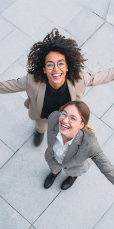Two cheerful multiethnic businesswomen standing together and looking up towards the sky, smiling happily with arms open, celebrating business success or a bright futureの素材
