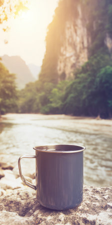 Metal mug of steaming hot coffee on a textured rock at sunrise, overlooking a tranquil river winding through lush karst hills-peaceful morning scene for outdoor adventure and calmの素材