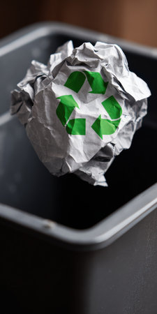 Crumpled paper with a prominent green recycling symbol is seen falling into an empty gray waste bin, symbolizing environmental consciousness, waste management, and the act of reusing materialsの素材