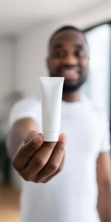 African american man is holding a white blank squeeze tube, presenting a cosmetic product for skin care or beauty, with an empty label for mockup copy spaceの素材