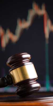 Gavel resting on a wooden desk with a blurry financial stock market chart displaying upward and downward trends in the background, representing legal oversight and economic policyの素材