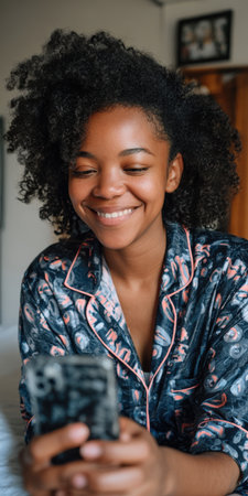 Young black woman with natural curly hair relaxing in pajamas at home, smiling as she looks at her smartphone while texting, browsing social media, and enjoying leisure timeの素材