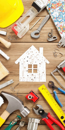 House shape cutout with weathered texture lying on a wooden background, surrounded by various home improvement and building tools, symbolizing renovation and diy projectsの素材