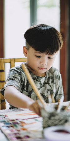 Young boy concentrating on watercolor painting at a home table, developing artistic skills and creativity while learning a new hobby with brush and palette, colorful close-upの素材