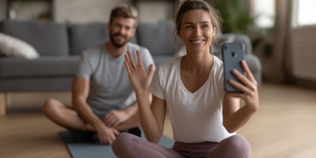 Smiling woman holding a smartphone, waving at the screen during a video call while exercising at home with her partner, connecting virtually for fitnessの素材