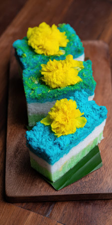 Three vibrant blue, white, and green layered steamed cakes, artistically arranged with yellow edible flowers and a banana leaf, resting on a rustic wooden platterの素材
