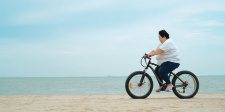 Overweight woman on a black fat bike riding along sandy beach by the sea, side view full body, active outdoor fitness, body positivity, joy and determination under blue skyの素材
