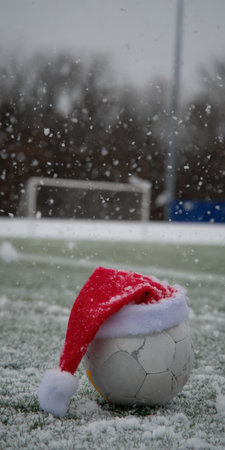 Soccer ball wearing a festive santa hat on a snow-covered artificial football field during a winter snowfall, combining holiday cheer with the love of sportの素材