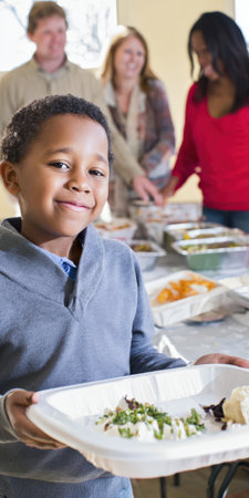 Young african american boy smiling and holding a disposable plate of food, standing in front of volunteers serving meals at a busy soup kitchen buffetの素材