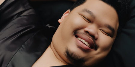 Asian man with beard relaxing on a couch, eyes closed and smiling widely, radiating genuine happiness, peaceful contentment and calm domestic wellbeingの素材