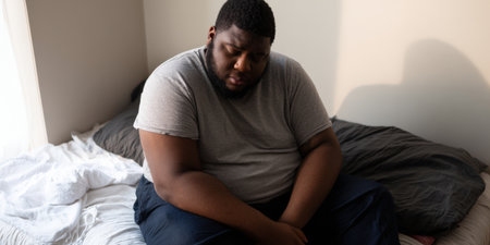 Overweight man sitting alone on a bed, looking down with a sad expression, conveying feelings of depression, loneliness, mental health issues, and deep worryの素材