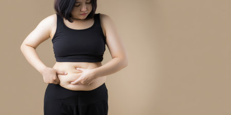 Woman standing in black sportswear, pinching her belly fat with both hands, looking down with a somber expression, expressing concepts of body shame, weight gain, insecurity, and health concernの素材