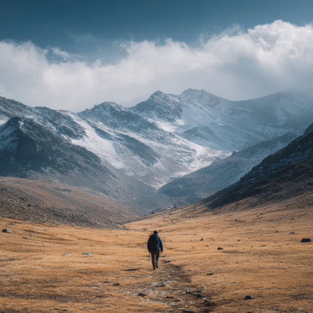 Man walking on a scenic path through a vast, golden mountain valley, surrounded by rugged snow-capped peaks under a cloudy sky, representing adventure, travel, and solitudeの素材