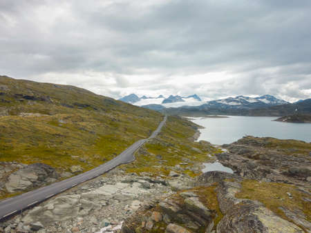 straight run through tundra of stones and grasses beside a lake towards the snow-capped mountains on a cloudy summer day in Jotunheimen National Park in Norway.の写真素材
