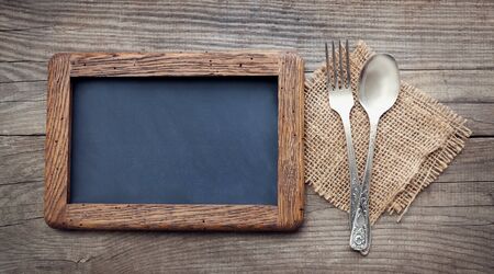 fork and spoon on a rustic table with a rough frame for chalk records, blackboard next to the dining utensils, top viewの写真素材