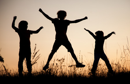 Children playing jumping on summer sunset meadow silhouetted.