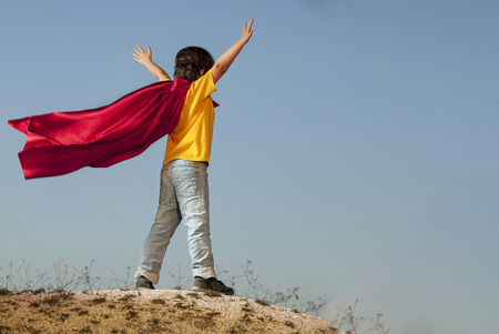 Boy playing superheroes on the sky background, teenage superhero in a red cloak on a hillの写真素材