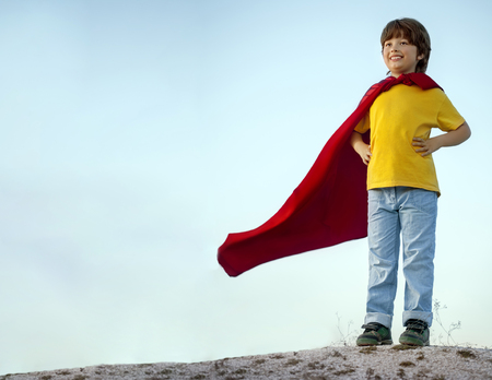 Boy playing superheroes on the sky background, child superhero in a red cloak on a hillの写真素材