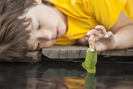 green leaf-ship in children hand in water, boy in park play with boat in river (focus on leaf)の写真素材