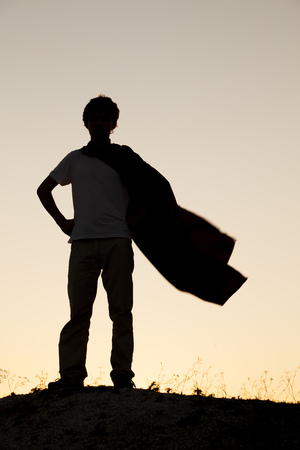 Boy playing superheroes on the sky background, teenage superhero in a red cloak on a hillの写真素材