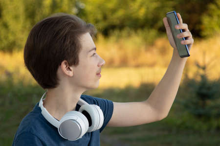 A teenager in a blue jacket with headphones and makes selfie on the green summer backgroundの写真素材