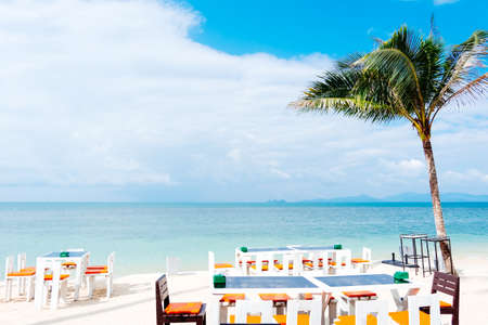 Outdoor white dinner table setting on white sand summer beach, coconut palm tree and calm blue sea...