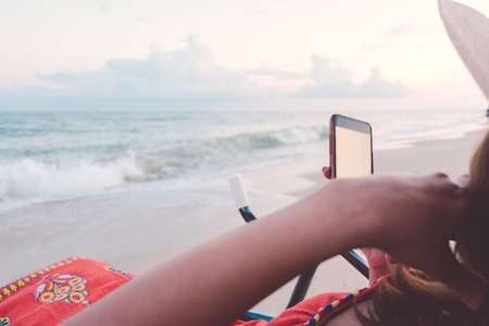 Woman in hat sitting on beach chair holding blank white screen smartphone taking photo, reading news, looking photo vacation on tropical beach with blurred summer sea. Lifestyle, Travel, Technology.の写真素材