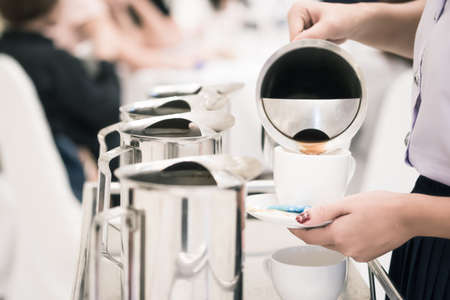 Close up woman staff is pouring coffee in pot to serve guests attending seminar during break time with blurred people in meeting room.の写真素材