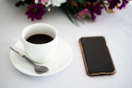 White cup of black coffee with blank smartphone screen and flower placed on white table background at coffee shop in fresh morning. の写真素材