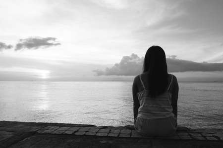 Silhouette of beautiful woman sitting alone on back side outdoor at tropical island beach missing boyfriend and family in summer sunset. Sad and lonely concept in black and white colour.の写真素材