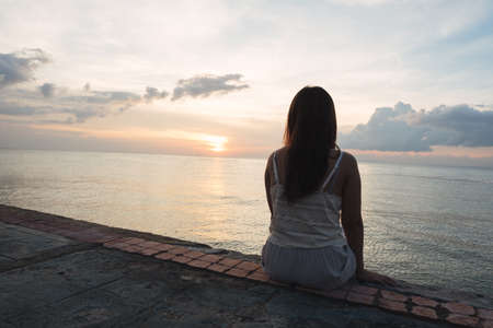 Silhouette of young woman sitting alone on back side outdoor at tropical island beach missing boyfriend and family in summer sunset. Sad and lonely concept in dark and vintage tone.の写真素材