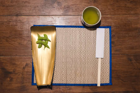 Wooden chopsticks in paper cover, boiled green soya bean and salt placed on gold plate on bamboo mat, cup of hot green tea on wooden table background. Top view Japanese food equipment and restaurant.の写真素材